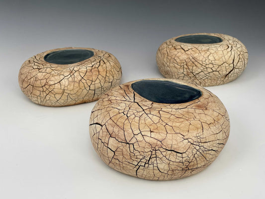 A group of three slightly flattened rounded ceramic forms with a  pool of blue glaze on the top near the center of each, surrounded by a tan matte surface with a dark brown cracked texture over each form.