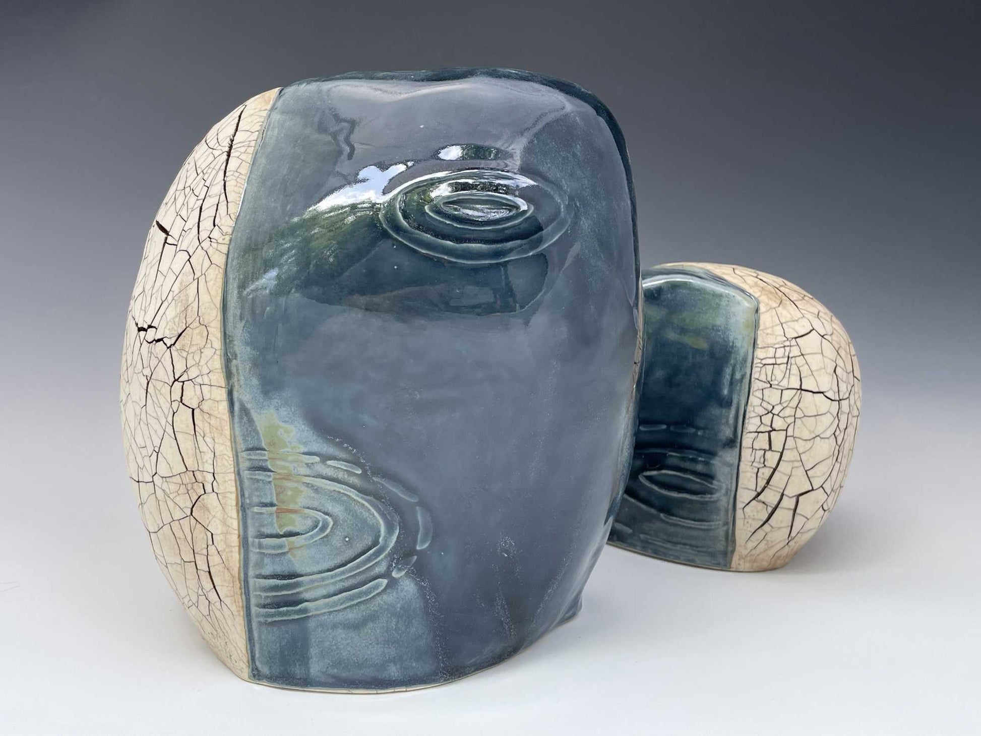 a sculpture consisting of a pair of oval ceramic forms.  Each has a dark blue glossy glaze over raindrop imagery on one side and a tan, matte cracked surface on the other side.