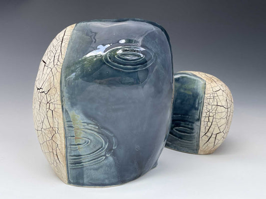 a sculpture consisting of a pair of oval ceramic forms.  Each has a dark blue glossy glaze over raindrop imagery on one side and a tan, matte cracked surface on the other side.