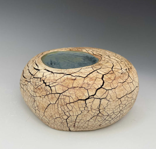 A slightly flattened rounded ceramic form with a  pool of blue glaze on the top near the center, surrounded by a tan matte surface with a dark brown cracked texture over the form.