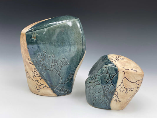 A pair of abstract organic-shaped ceramic sculptural forms.  Each has fine dark brown branching lines, representing seaweed flowing over the forms. Surfaces are finished with each having half the piece covered in a dark blue glossy glaze and half a tan, matte surface.