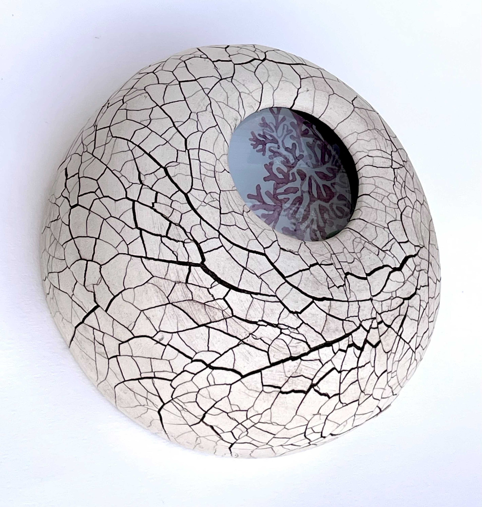 wall-hung sculpture, a round ceramic form with a tan matte sruface with a dark brown crackle texture.  A painting of pink seaweed is visible through an inset small round glass lens near the center of the piece