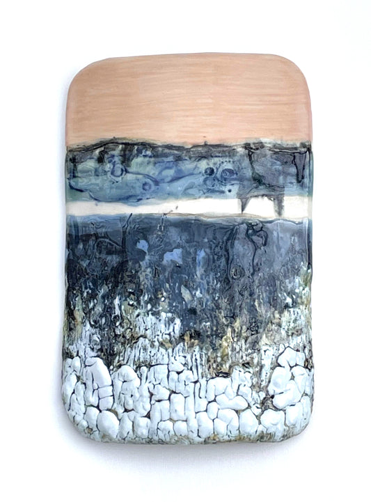 Rectangular porcelain wall-hung sculptural tile with coral, white, blue and dark grey glazes on a textured surface, representing an abstract sunset landscape scene